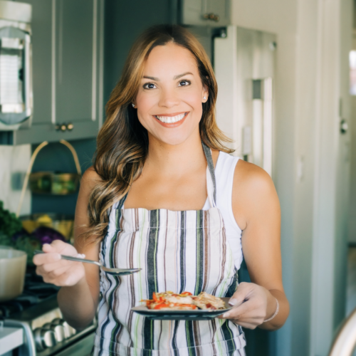 A smiling woman in a kitchen holds a plate with food. She wears a striped apron and holds a spoon. The kitchen has visible cabinets, and there's a basket with vegetables in the background.