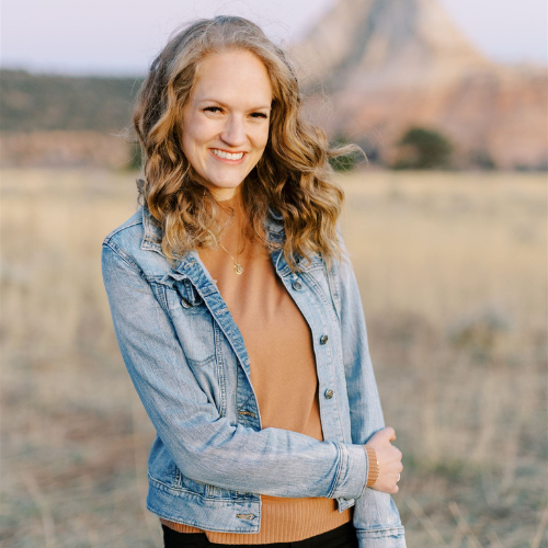 A woman with wavy blonde hair smiles warmly at the camera. She is wearing a light blue denim jacket over a brown top and stands in front of a scenic, blurred background featuring a field and a rocky hill or mountain.