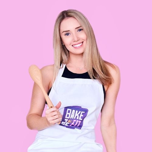 A smiling woman with long blonde hair holds a wooden spoon. She wears a white apron with a "Bake Off" logo on a pink background.