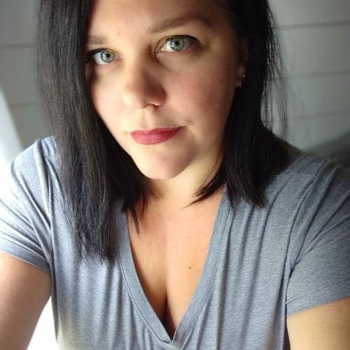 The image shows a person with shoulder-length black hair and a neutral expression, wearing a gray V-neck shirt. The background is blurred, suggesting indoor lighting.