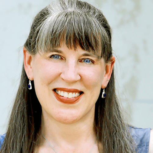 A woman with straight, shoulder-length gray hair, bangs, and blue eyes is smiling. She is wearing drop earrings and a necklace. The background is blurred and light-colored.