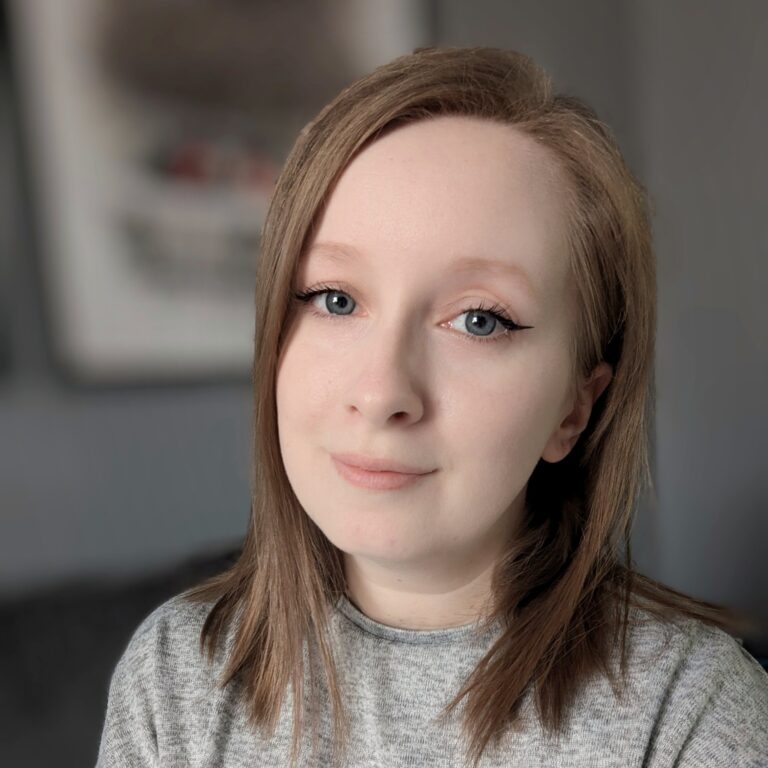 A woman with straight, light brown hair and blue eyes is wearing a gray sweater. She has subtle makeup with winged eyeliner. The background is blurred, featuring a grayish wall with a framed picture.