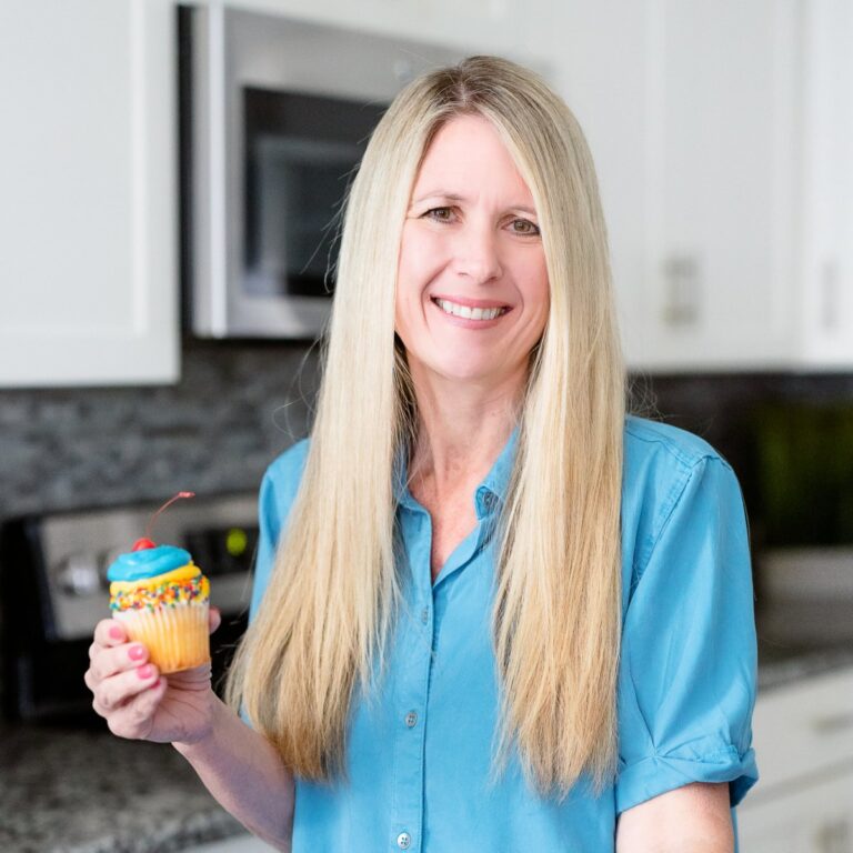 A person with long blonde hair is smiling and holding a cupcake with blue frosting and a cherry on top, standing in a kitchen with a stove and cabinets in the background. They are wearing a blue shirt.