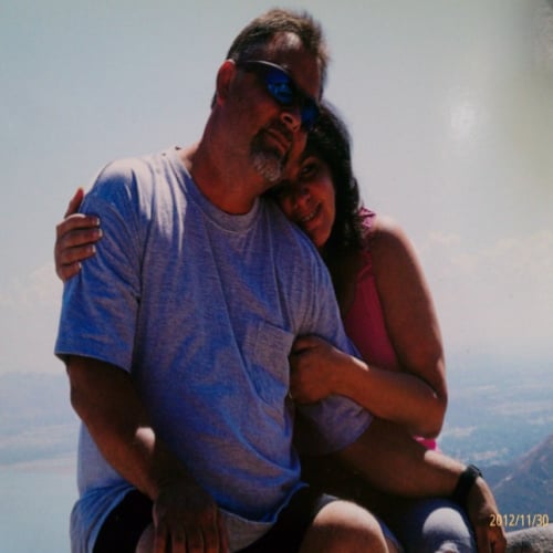 A man in a gray T-shirt and sunglasses sits with a woman in a pink top, embracing him. They are outdoors with a hazy view of water and hills in the background. The photo appears sunlit, and they are smiling.