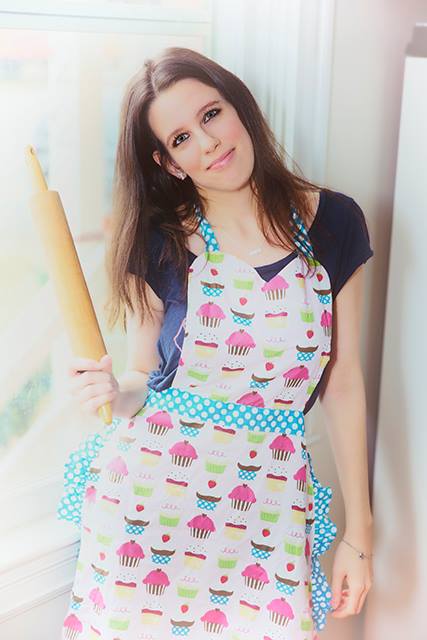 A person with long hair holds a rolling pin while wearing a colorful apron with cupcake prints. They are standing near a window, smiling.