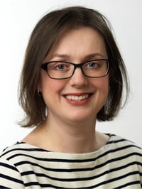 A woman with shoulder-length brown hair and glasses smiles at the camera. She is wearing a white shirt with black stripes against a plain white background.