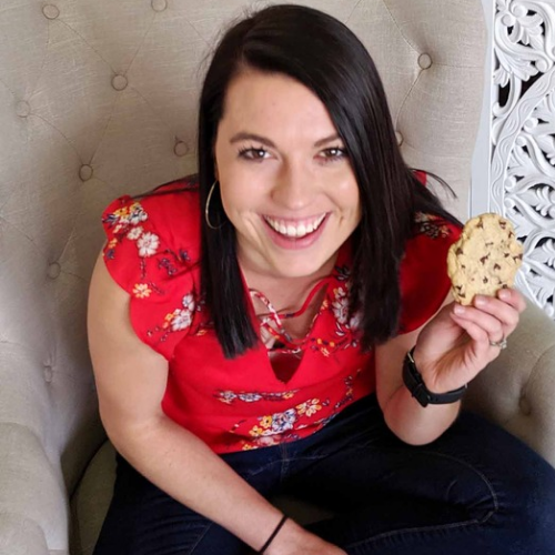 A woman with dark hair, wearing a red floral blouse, is seated on a light-colored cushioned chair. She is smiling broadly and holding a large, round cookie in her right hand. The background features a decorative white design on the right side.
