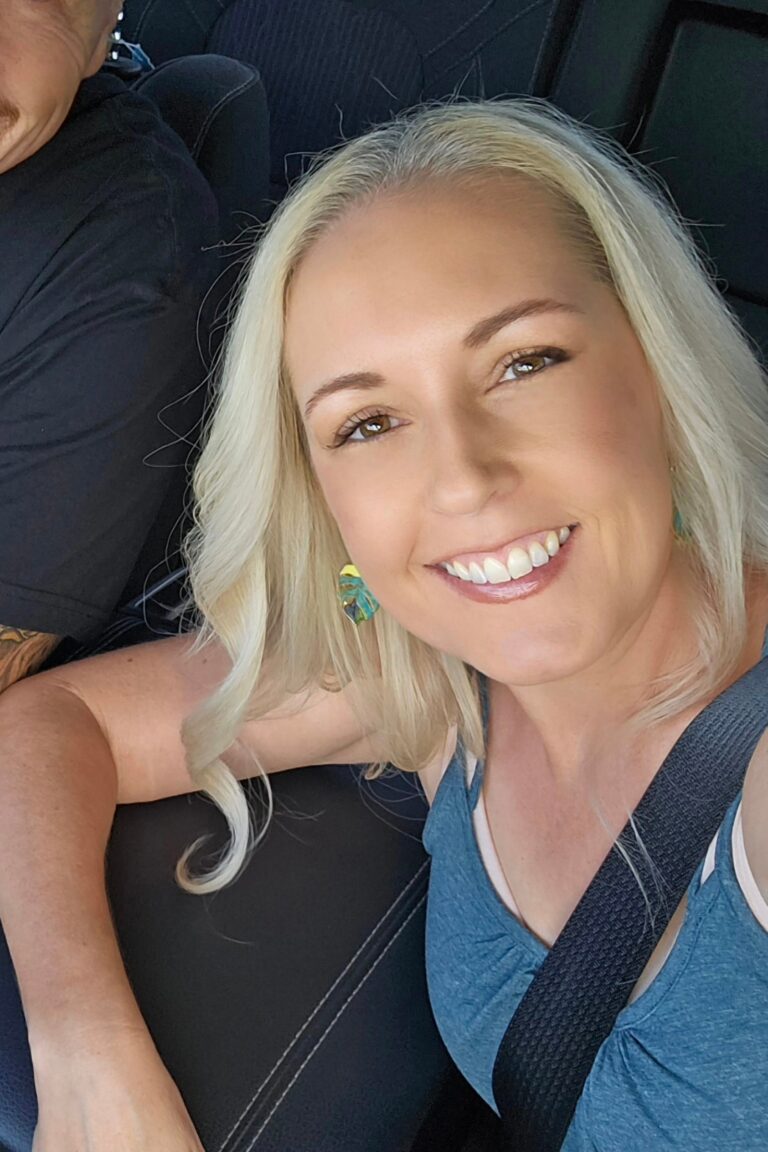 Smiling woman with long blonde hair taking a selfie inside a car. She's wearing a blue top and colorful earrings, with a seatbelt fastened. The edge of another person is visible on the left.