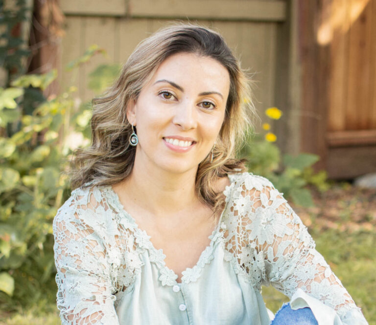 A woman with wavy, shoulder-length hair smiles at the camera while sitting outdoors. She is wearing a light blue blouse with lace detailing. The background features greenery and a wooden fence, evoking a sense of thriving in a natural, sunny ambiance.