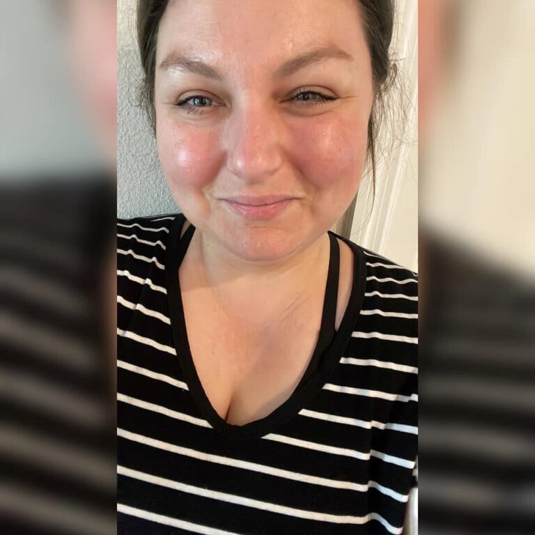 A person smiling softly at the camera, wearing a black and white striped shirt with a black neckline. Their hair is pulled back, and the background is softly blurred.