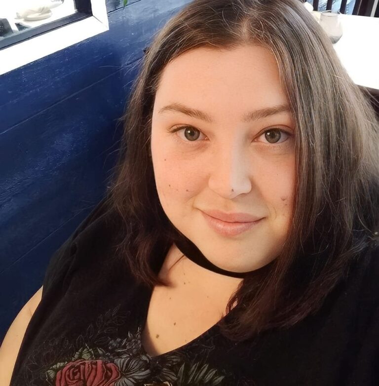 A person with shoulder-length brown hair is smiling at the camera. They are wearing a dark top with a subtle floral design, sitting in a room with blue walls and bright lighting.