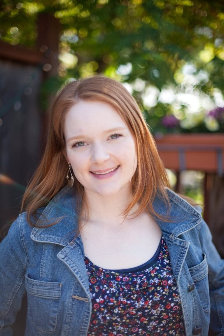 A person with long red hair smiles outdoors, wearing a denim jacket over a floral top. The background is sunny and blurred, featuring green foliage and some flowers.
