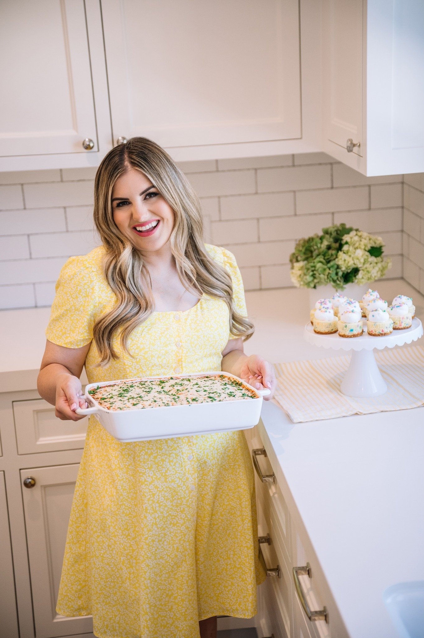 Woman in a yellow dress holding a casserole dish in a white kitchen. Cupcakes with frosting are on a stand nearby. There's a bouquet of white flowers on the counter.