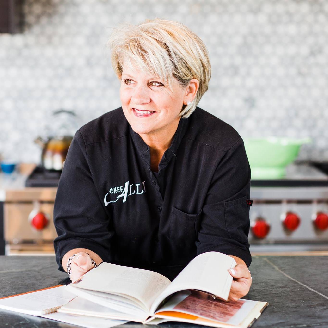 A woman with short blonde hair wearing a black chef jacket stands in a kitchen with a hexagon-tiled wall. She is holding an open cookbook and smiling, with a stove featuring red knobs visible in the background.
