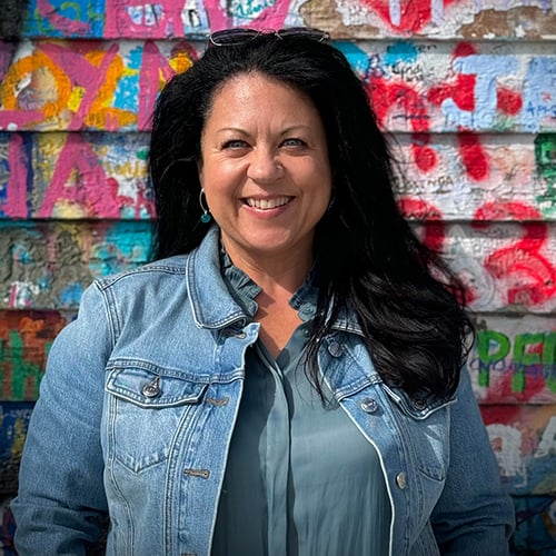 A person with long dark hair smiles while standing in front of a brightly colored, graffiti-covered wall. They are wearing a denim jacket and a gray shirt.