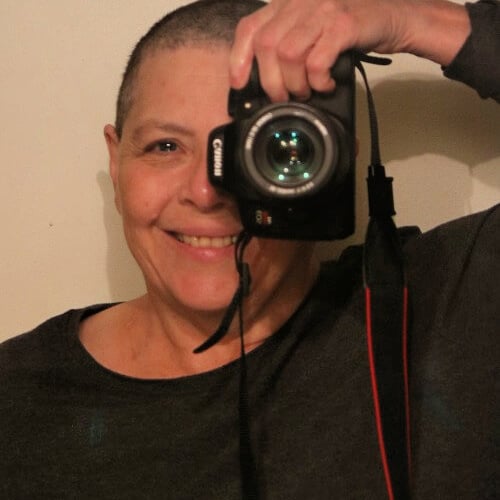 Person with short hair holds a camera up to their face, smiling. They are wearing a dark top, and the camera has a visible strap. The background is plain and neutral.