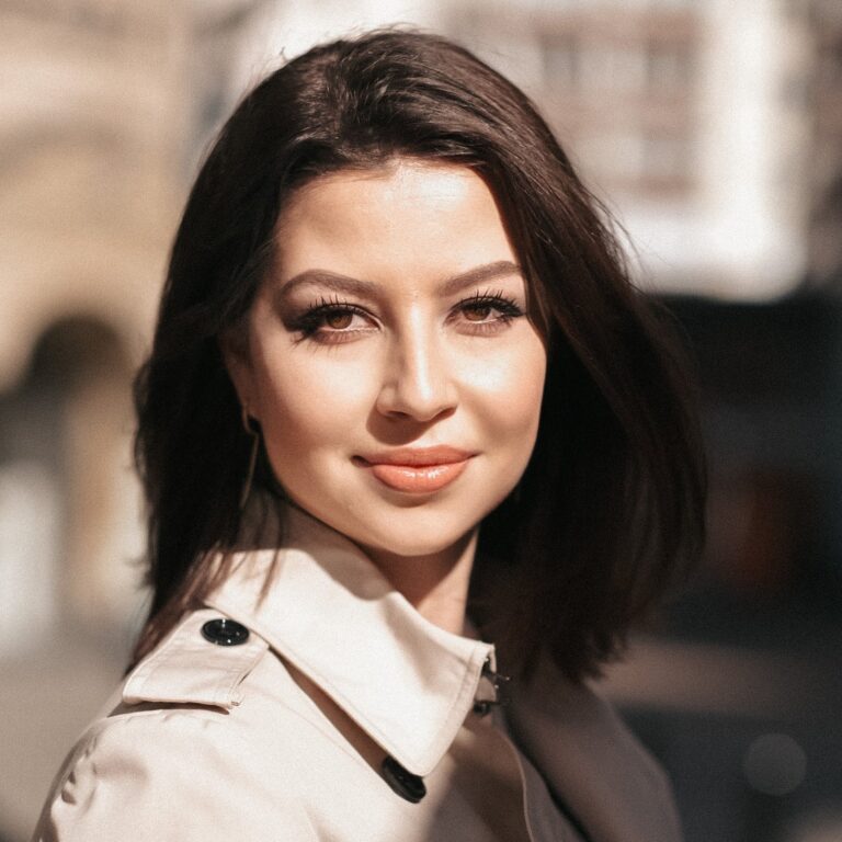 Woman with dark hair and a beige coat smiles outdoors under sunlight.