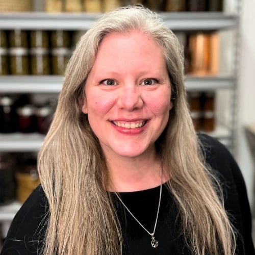 Smiling woman with long gray hair wearing a black top and a necklace. She is standing indoors with shelves filled with various items in the blurred background.