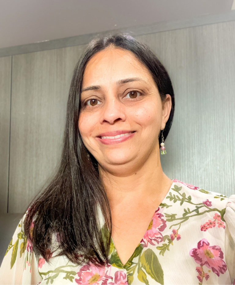 Smiling woman with long dark hair wearing a light floral blouse stands in front of a gray background.