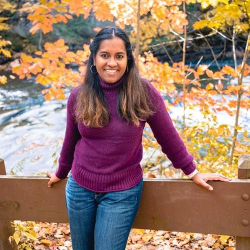 A person in a purple sweater and jeans smiles while leaning against a wooden railing. The background features a scenic view of autumn trees with orange leaves and a flowing river.