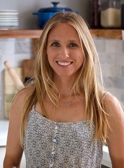 A woman with long blonde hair smiles at the camera. She is wearing a light patterned top and standing in a kitchen with a marble backsplash, wooden shelves, and various kitchen items in the background.