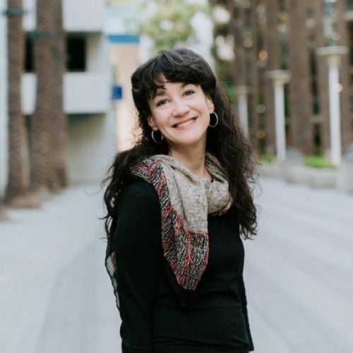 A woman with long dark hair smiles while standing outdoors. She is wearing a black top and a scarf. The background features a pathway lined with tall trees and a building.