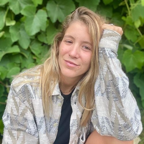 Young person with long blonde hair, wearing a patterned shirt over a black top, smiling with one hand on their head. Green leaves in the background.