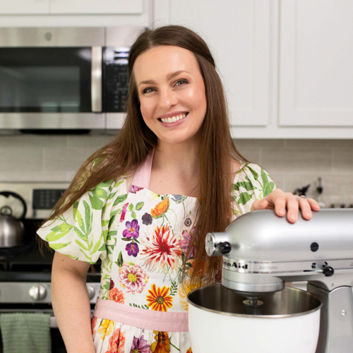 A smiling woman in a floral apron stands in a modern kitchen with her hand on a silver stand mixer. White cabinets, a microwave, and a kettle are visible in the background.