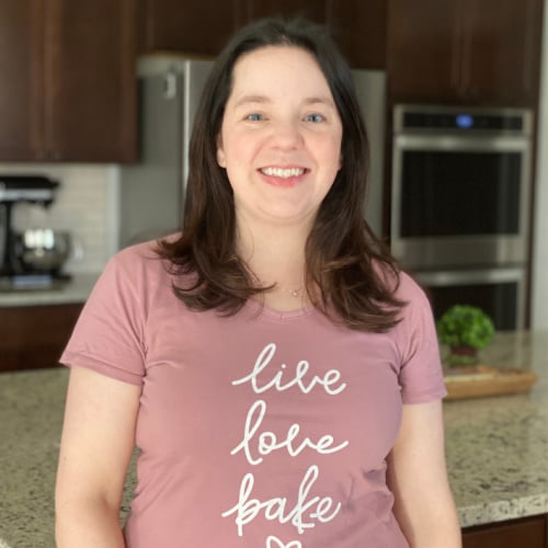A woman with long dark hair stands in a kitchen, smiling. She is wearing a pink shirt with the words "live love bake" on it. A plant and modern kitchen appliances are visible in the background.