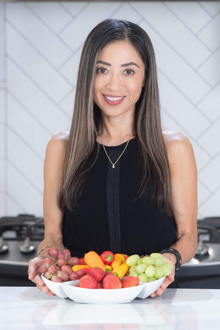 A woman with long brown hair wearing a black sleeveless top stands in a kitchen, smiling and holding a white bowl filled with grapes, strawberries, and mini bell peppers.