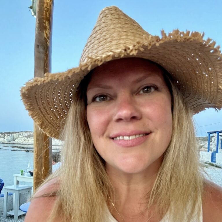 A woman smiles at the camera while wearing a straw hat. She has blonde hair and is outdoors near a beach setting with tables and chairs visible in the background, along with a clear blue sky and water.