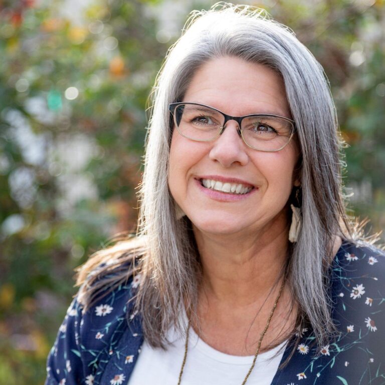 A woman with long gray hair and glasses is smiling outdoors. She is wearing a floral-patterned top over a white shirt. The background is blurred with greenery, suggesting a sunny day.