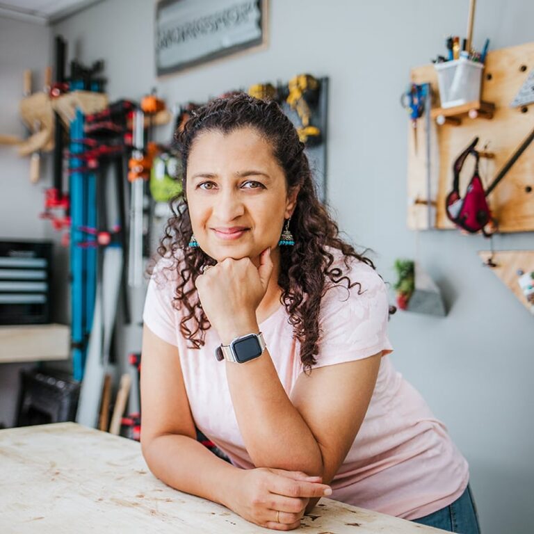 A person with curly hair stands at a wooden workbench in a workshop, resting their chin on their hand. They wear a pink top and a smartwatch. The background features tools and wooden shelves on the wall.