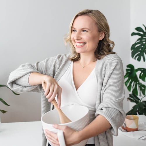 A smiling woman with blonde hair is happily mixing ingredients in a white bowl with a wooden spoon. She is wearing a light grey cardigan over a white top. There are green plants in the background.