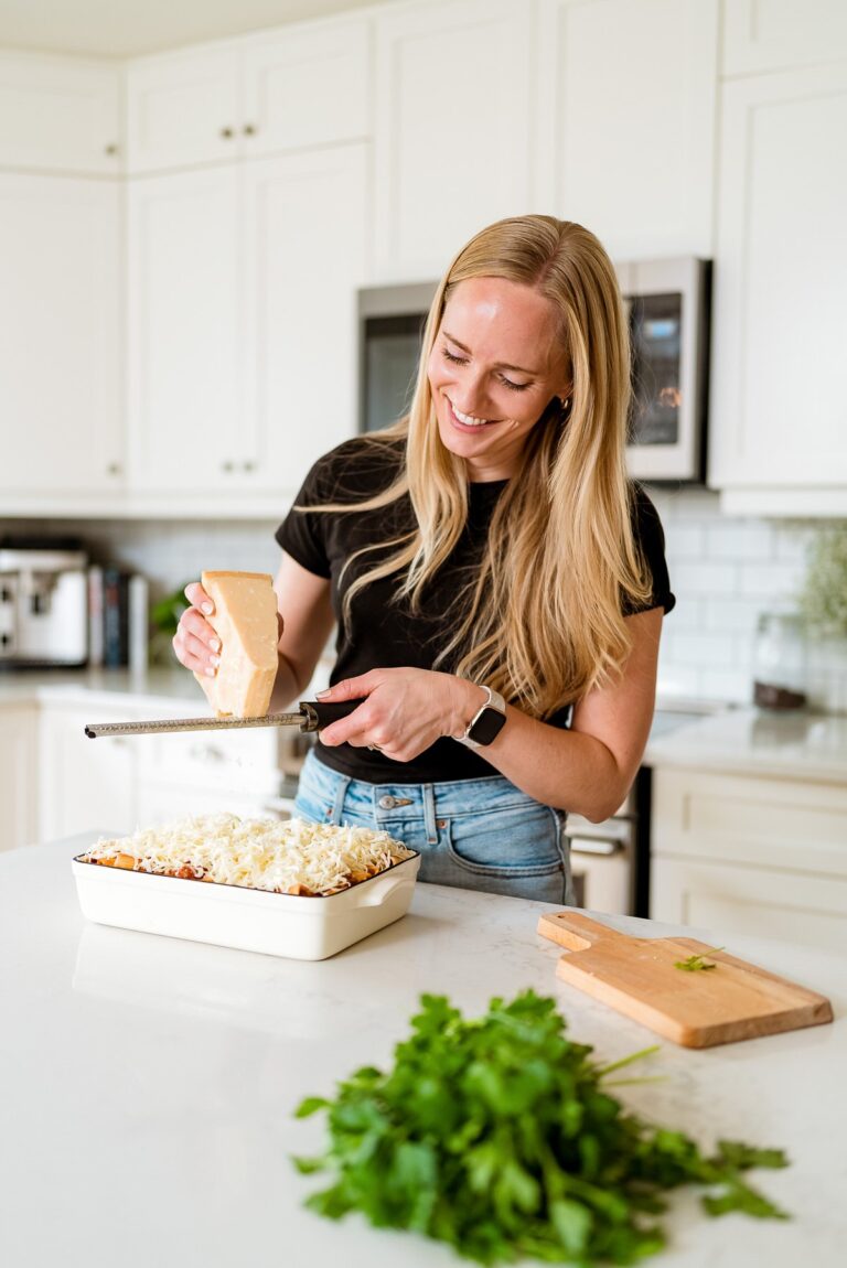 A woman with long blonde hair grates cheese over a dish in a bright kitchen. She wears a black shirt and jeans, smiling while preparing the meal. Fresh herbs and a cutting board are on the white countertop.