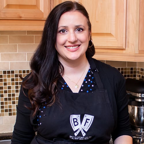 Woman with long dark hair smiling in a kitchen, wearing a black apron over a blue polka dot top. She stands in front of tiled backsplash and wood cabinets. A mixer is partially visible in the background.