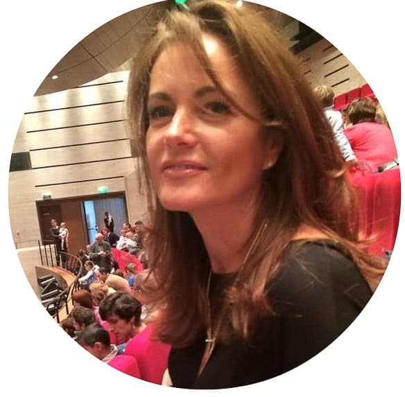 A woman with shoulder-length brown hair wearing a black top sits in an auditorium filled with people. She looks towards the camera with a slight smile. The room has red seats and a modern design, with people talking and waiting in the background.