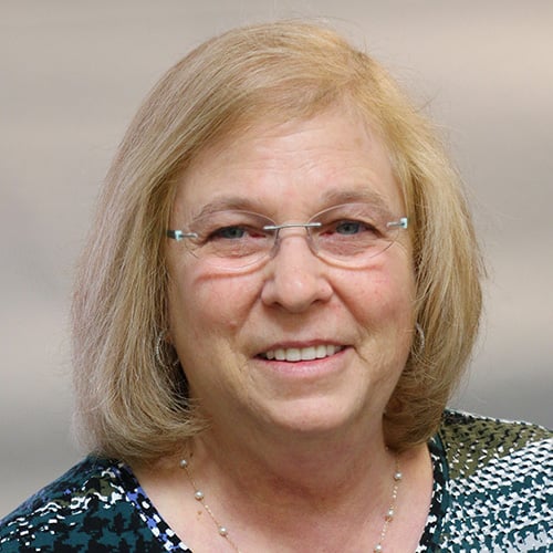 A woman with shoulder-length blond hair and glasses smiles at the camera. She is wearing a patterned top and a necklace. The background is a soft, neutral blur.