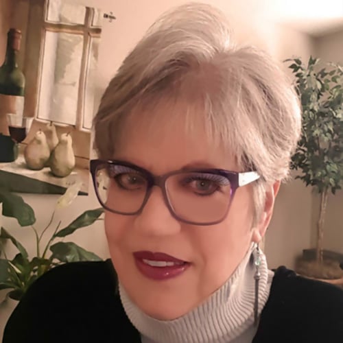 A person with short gray hair and glasses smiles at the camera. They are wearing a turtleneck sweater and earrings. In the background, there are plants and artwork on the wall.