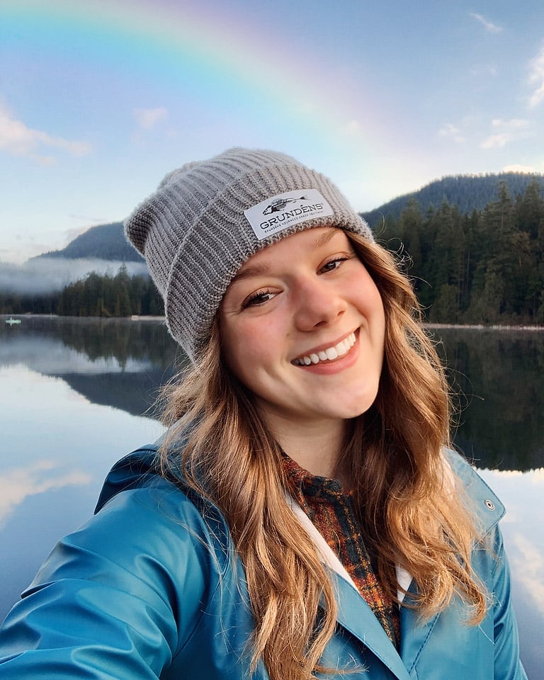 A person wearing a gray beanie and blue jacket smiles for a selfie. In the background, a serene lake reflects the surrounding forest and hills, with a vibrant rainbow arching across a clear sky.