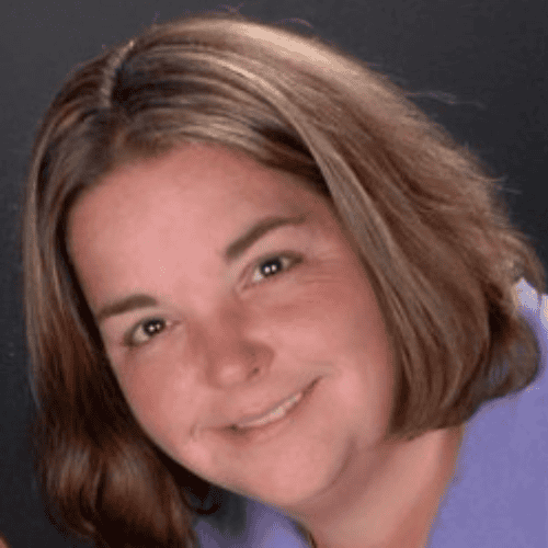 A woman with shoulder-length brown hair smiles at the camera. She is wearing a light purple top and has a relaxed, friendly expression on her face. The background is dark, bringing focus to her face.