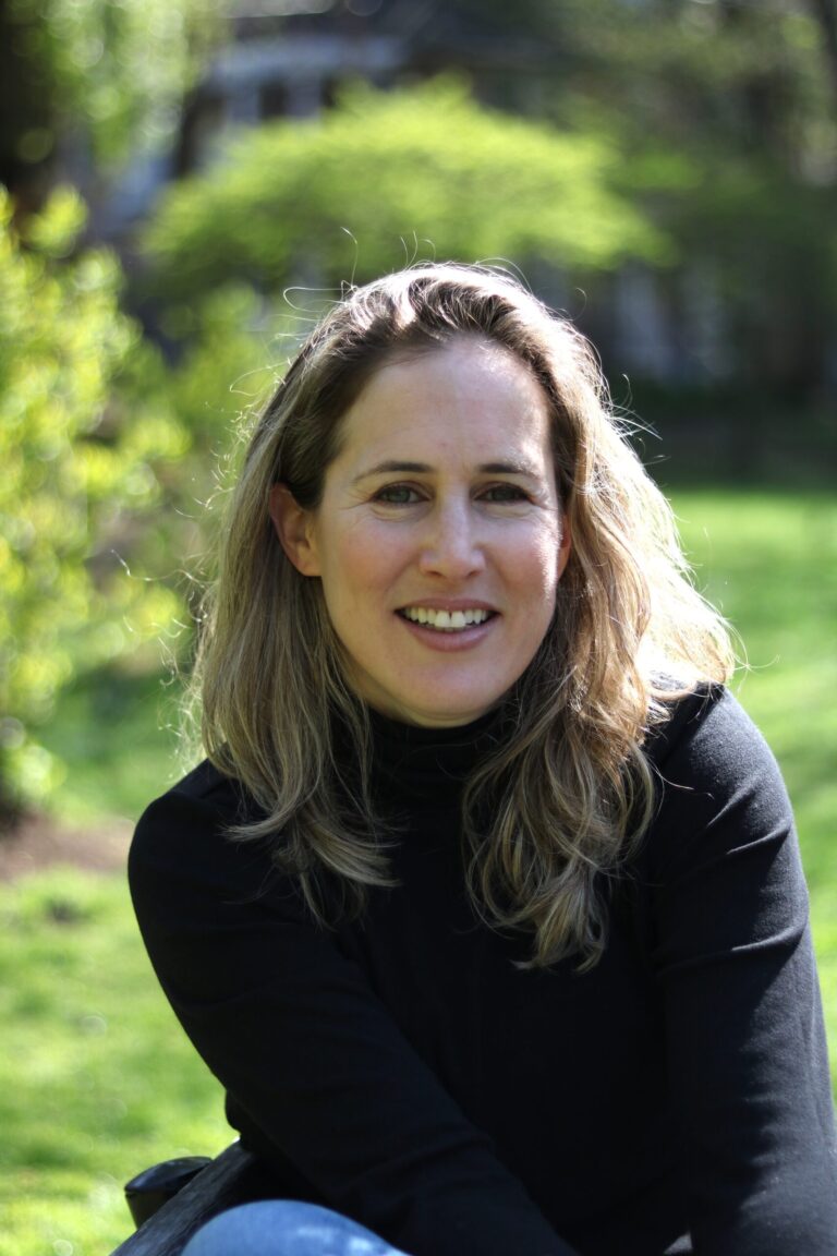 A woman with long blonde hair smiles while sitting outdoors. She is wearing a black top and is surrounded by greenery, with a blurred background of trees and grass.