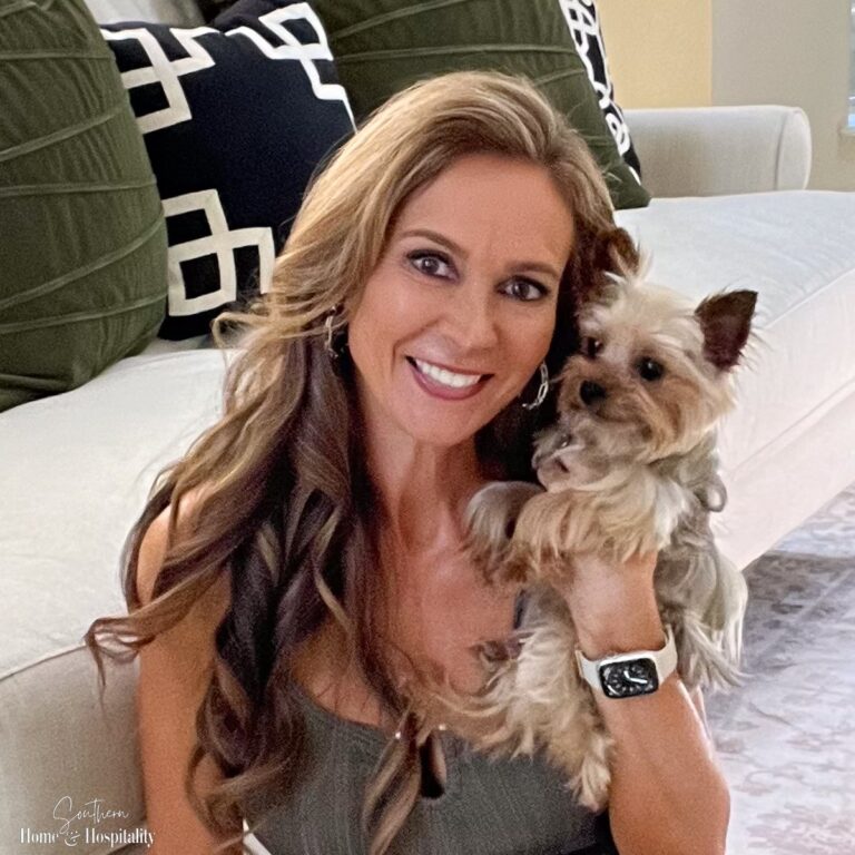 A woman with long brown hair smiles while holding a small Yorkshire Terrier. She is sitting on a couch with black and white patterned pillows.