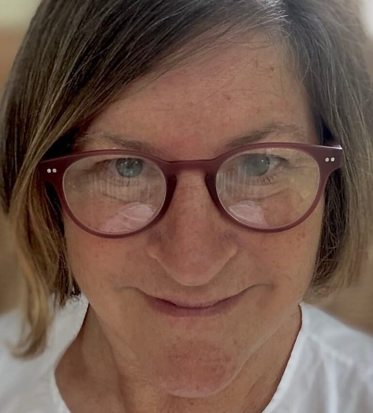 A close-up of a person with short brown hair wearing red glasses and a white shirt. They are looking directly at the camera and smiling slightly.