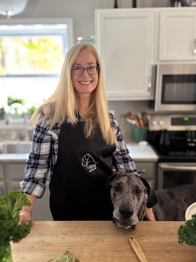 A woman with long blonde hair and glasses smiles in a kitchen while wearing a black apron with a dog logo. A large, friendly-looking dog rests its head on the countertop beside her. The kitchen features white cabinets and stainless steel appliances.