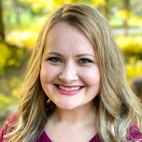 A person with long blonde hair smiles warmly at the camera. They are wearing a red top. The background is a softly focused natural setting with green foliage and sunlight filtering through.