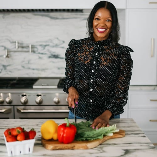 A woman in a black, polka-dotted blouse smiles while slicing a red bell pepper on a wooden board in a modern kitchen. Other vegetables and a container of strawberries are on the counter.