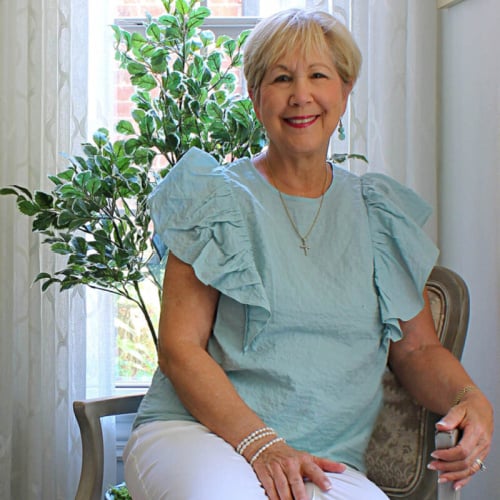 A woman with short blonde hair sits on a chair indoors, smiling. She wears a light blue blouse with ruffled sleeves and white pants. A leafy plant and sheer curtains are in the background.