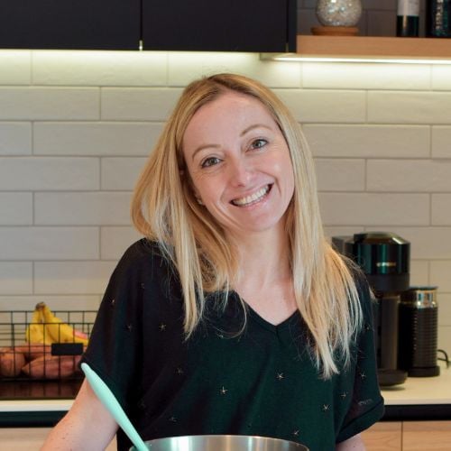 Smiling person with long blonde hair in a kitchen, wearing a black shirt. They stand in front of a gray tiled wall with a bowl and utensils. Various kitchen items, including a fruit basket and a coffee maker, are visible in the background.