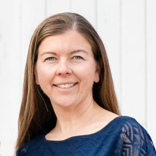 A woman with long, straight brown hair smiles while wearing a dark blue patterned blouse. She stands against a white background.